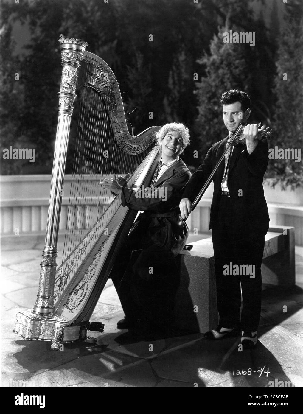 HARPO MARX plays his harp accompanied by violinist on set candid at Paramount Astoria Studios in New York during filming of ANIMAL CRACKERS 1930 director VICTOR HEERMAN based on musical play by George S. Kaufman Morrie Ryskind Bert Kalmar and Harry Ruby Paramount Pictures Stock Photo