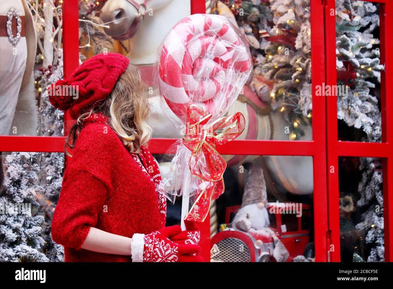 Girl in a red sweater with lollipop enjoys the magic of Christmas holidays standing on a street at a toy store window Stock Photo