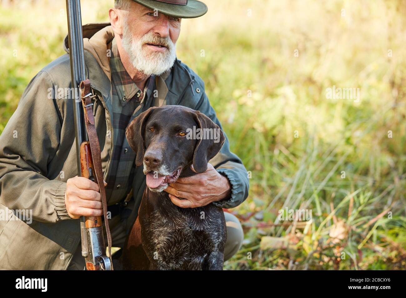 Senior man hug his hunter dog in autumn forest. Man wearing casual hunting clothes, sitting on ground.Gun in hands Stock Photo