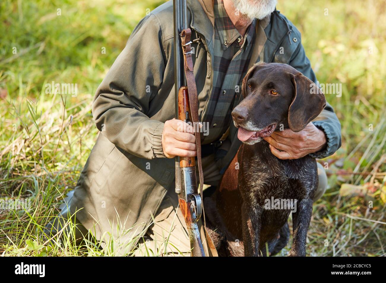 Cropped man with beard hug his hunter dog in autumn forest. Man wearing casual hunting clothes, sitting on ground.Gun in hands Stock Photo