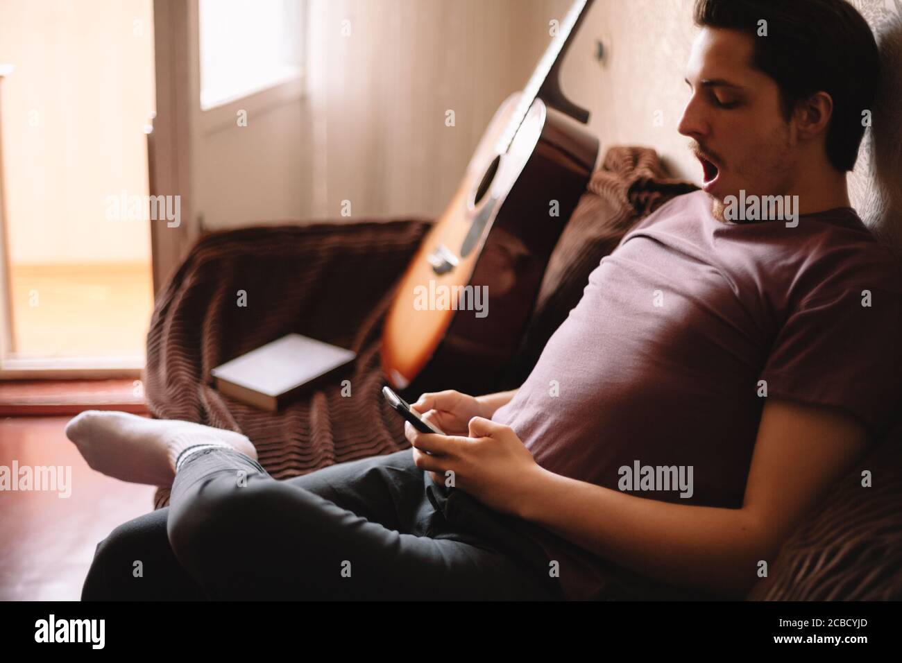 Young man yawning while using smart phone sitting at home Stock Photo