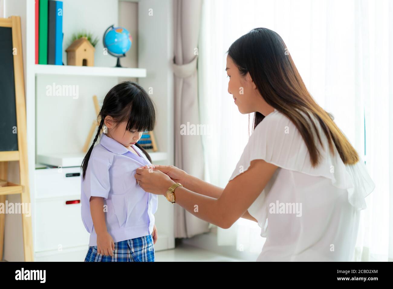 Asian mother preparing kindergarten student uniform to her little daughter for school getting ready for school in living room at home. Parenthood or l Stock Photo