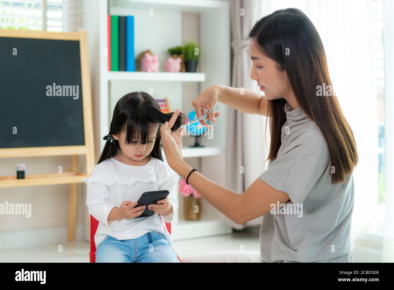 Asian Mother cutting hair to her daughter in living room at home while stay at home safe from Covid-19 Coronavirus during lockdown. Self-quarantine an Stock Photo