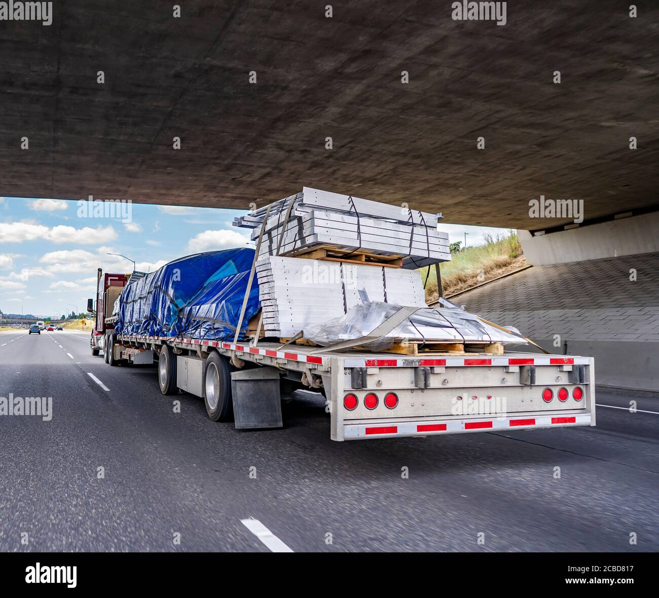 Hard loaded classic red big rig semi truck transporting covered and tightened commercial cargo on flat bed semi trailer running under the bridge acros Stock Photo