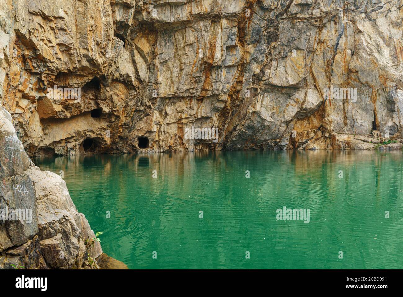 A green-colored lake and a rocky cliff with holes in abandoned adits. Tuim is a sinkhole at the site of an abandoned copper and tungsten ore mine. Stock Photo