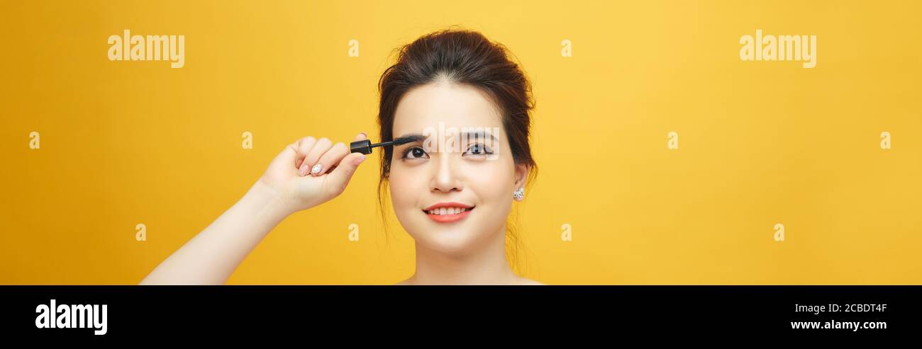 Closeup portrait of attractive young woman putting some mascara onto her eyelashes with make up brush over yellow background Stock Photo