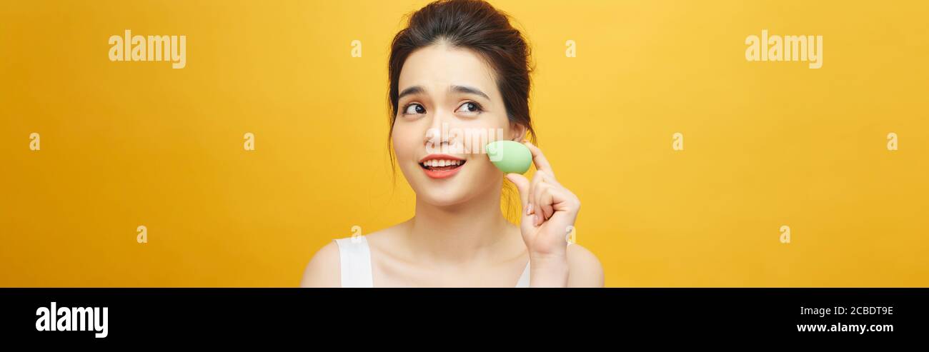 Beautiful young woman applying makeup using beauty blender sponge. Isolated over yellow background Stock Photo