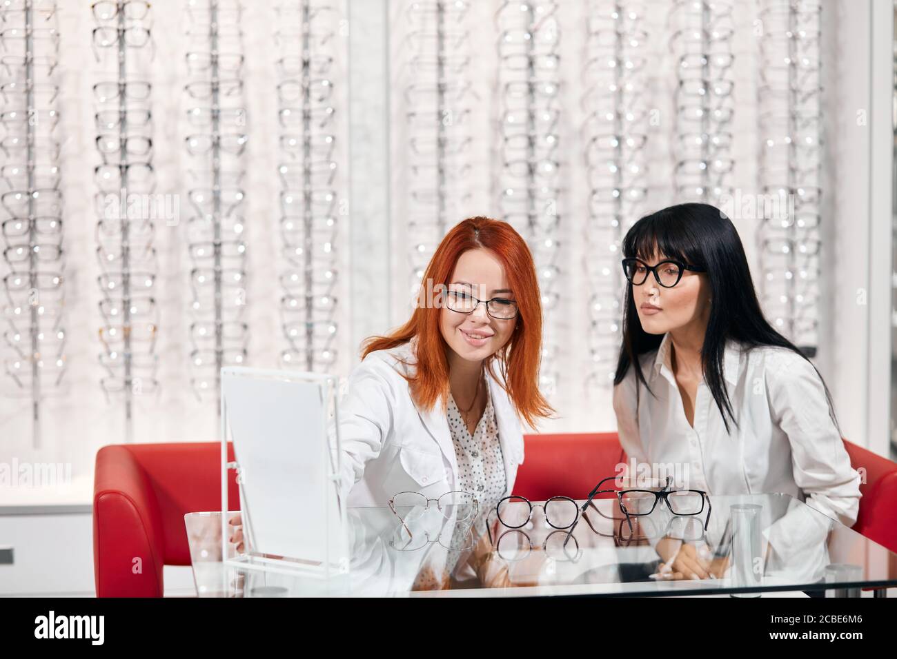 two beautiful optician sitting at the table and testing new eyeglasses at optics store Stock Photo