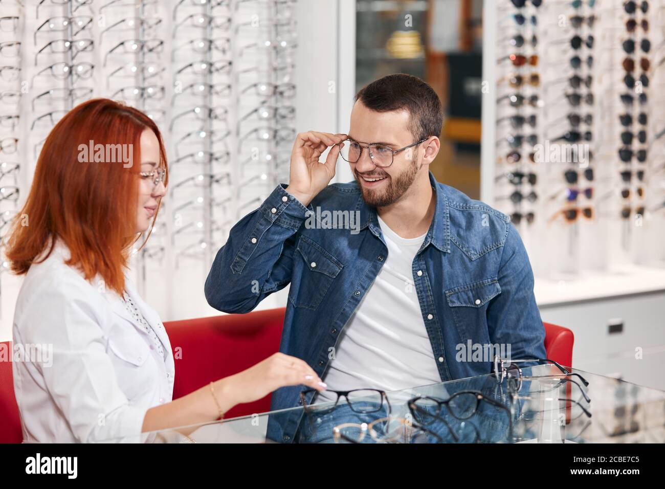 young positive man in denim jacket trying on different many glasses, close up photo. glasses on the shelf in the background of the photo Stock Photo