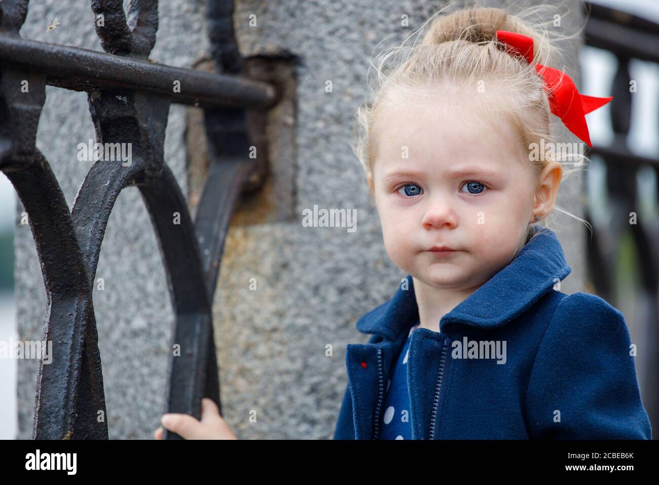 sad little girl in a blue coat and a red bow on a city street Stock Photo