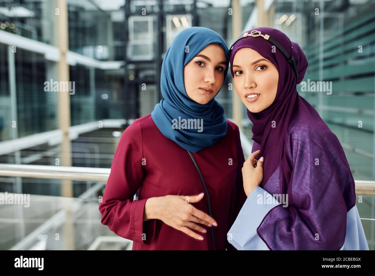 Couple of muslim caucasian 20s girlfriends with hijab or islamic head scarfs and islamic traditional clothes looking at camera standing in modern glas Stock Photo