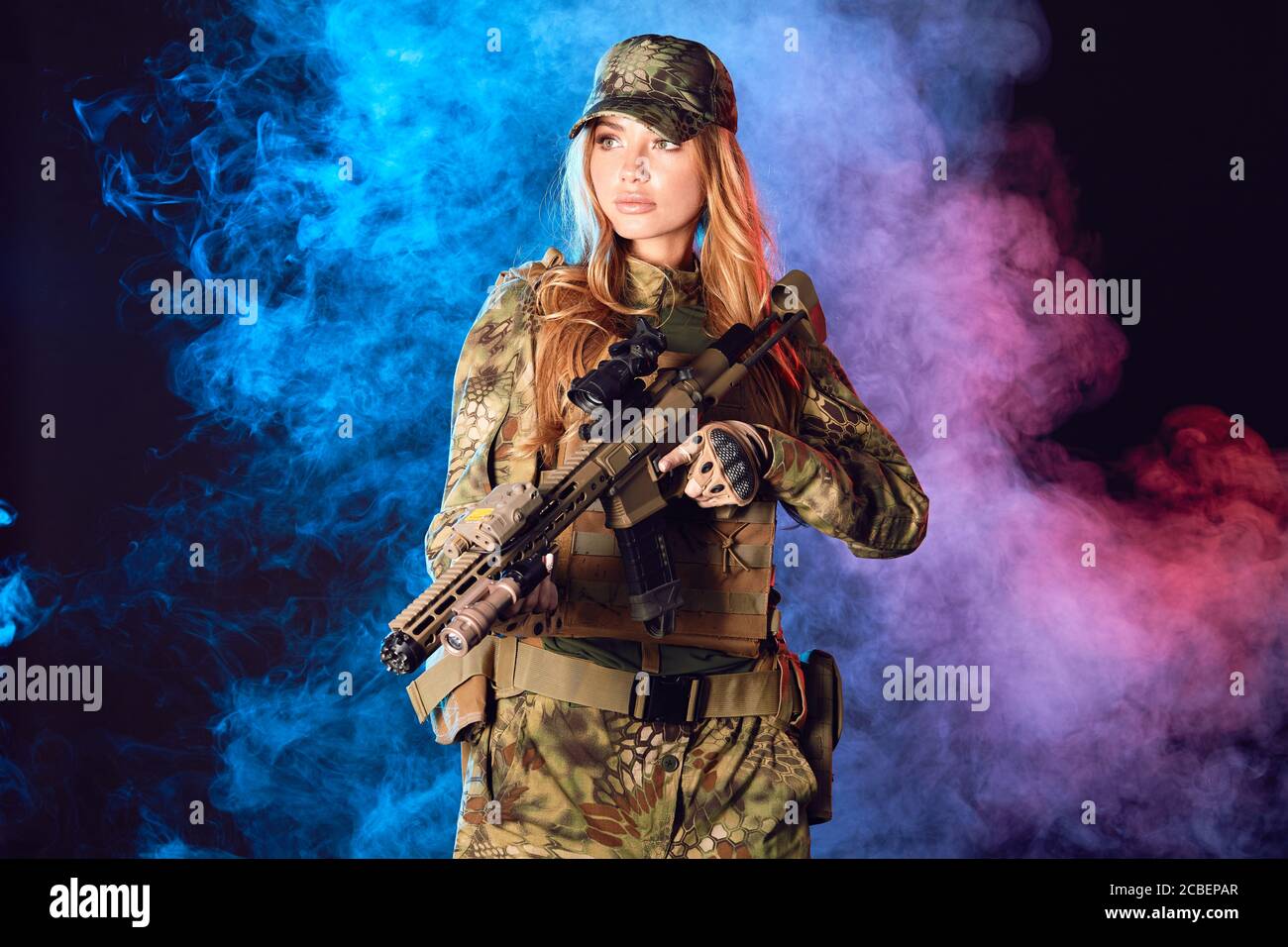 Female soldier in military camouflage uniform and cap holding sniper rifle over black background with smoky clouds Stock Photo