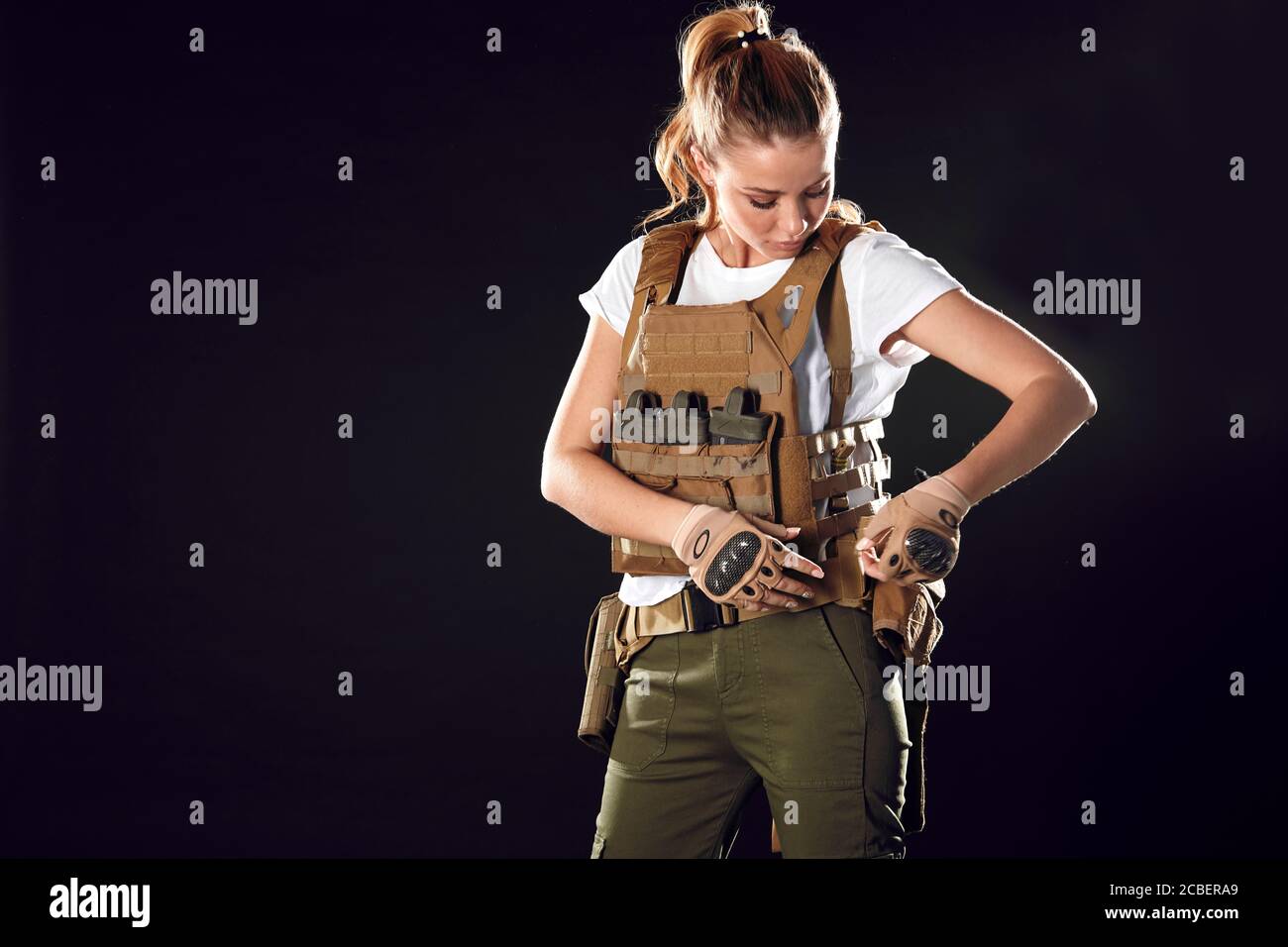 Gorgeous young woman with long blonde hair wearing Military gear, posing isolated in studio over dark background Stock Photo