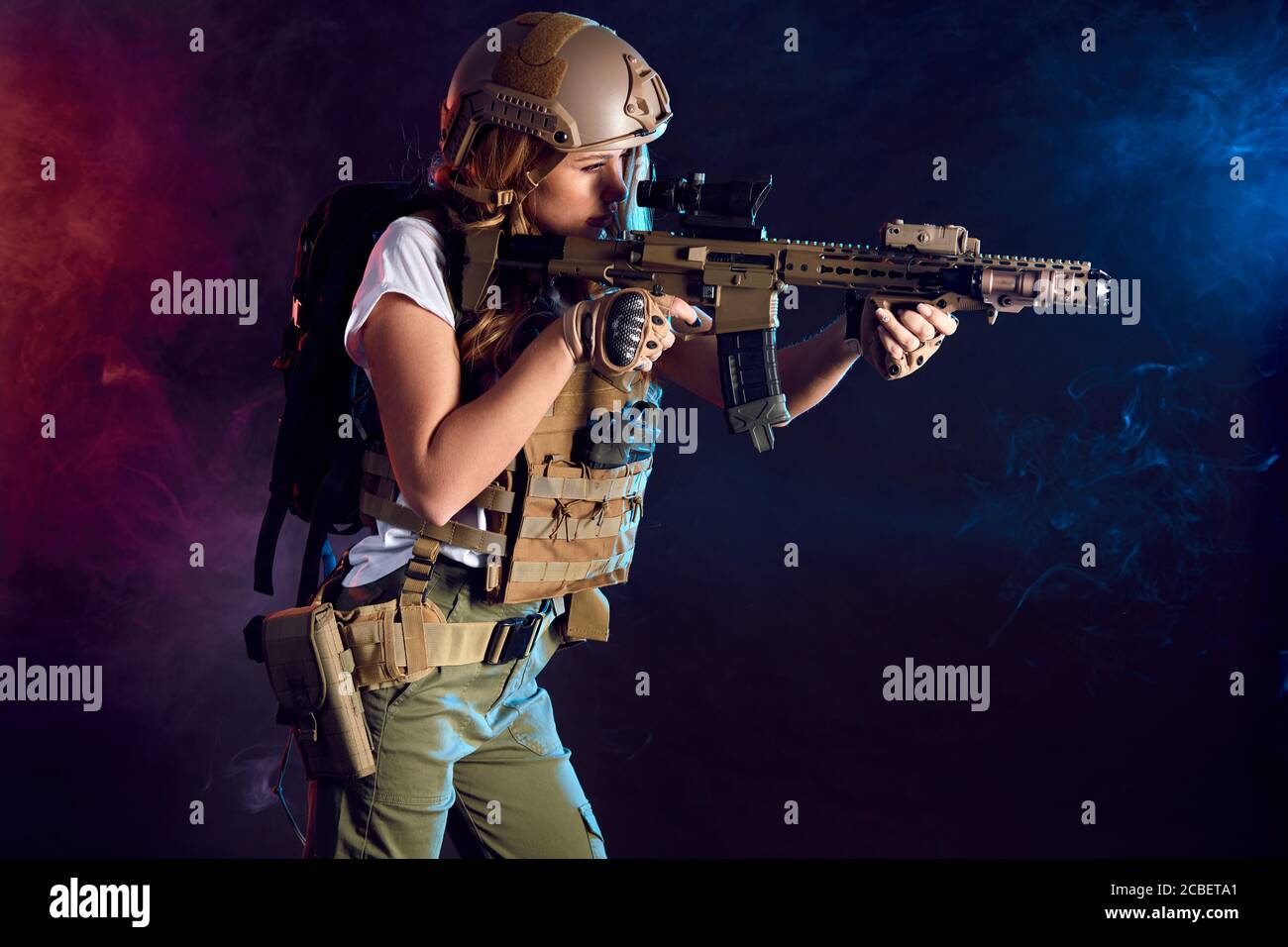 Heavily armed and well-equipped female soldier in battle helmet holding assault rifle isolated on dark smoky battlefield. Paint ball and laser tag spo Stock Photo