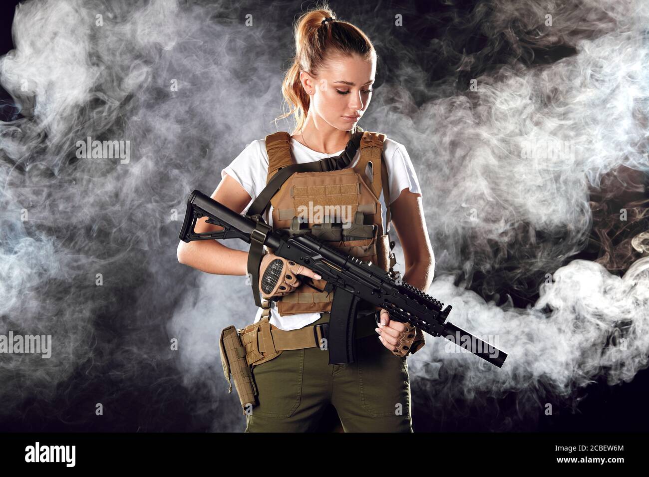Young blonde female snipper in military outfit with assault rifle in studio on smoky dark background. Women in military service concept. Stock Photo