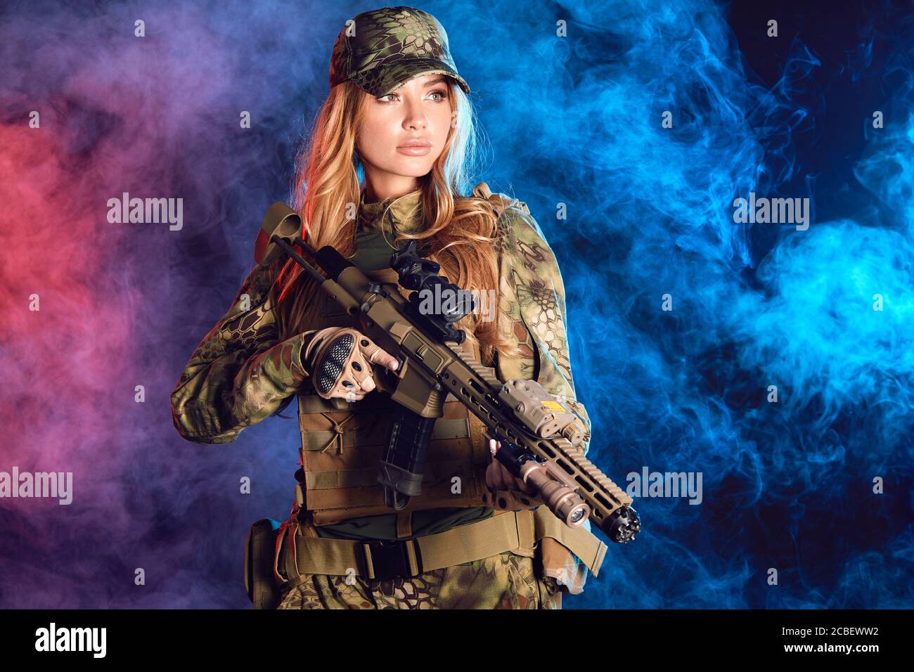 Female soldier in military camouflage uniform and cap holding sniper rifle over black background with smoky clouds Stock Photo