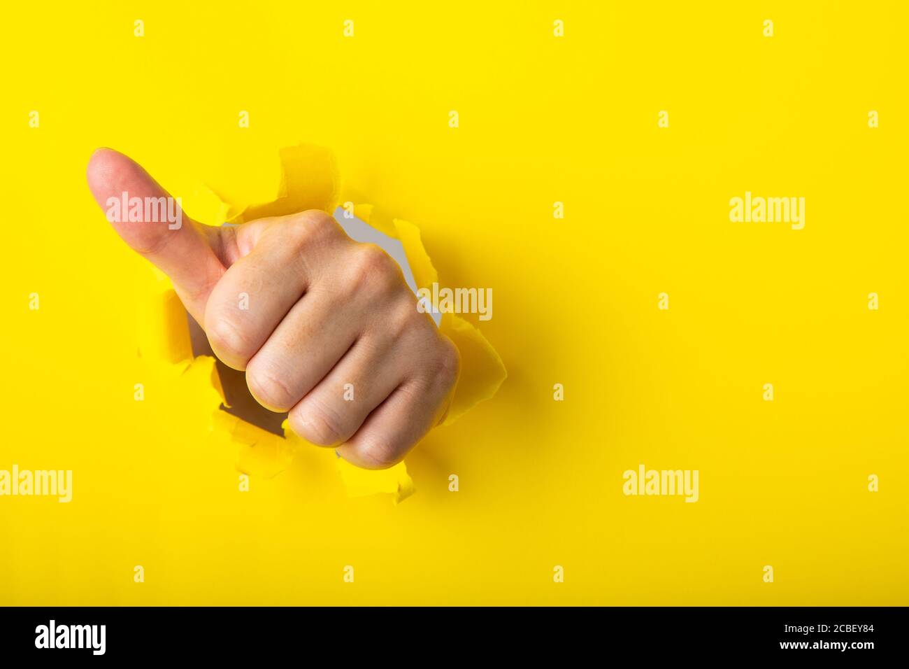 Hand showing a thumb up sign through a ripped hole in a yellow paper ...