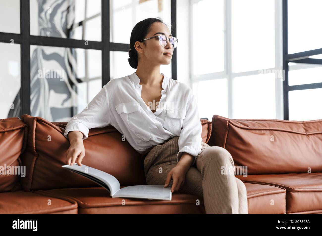 Attractive smiling young asian business woman relaxing on a leather couch at home, reading a magazine Stock Photo