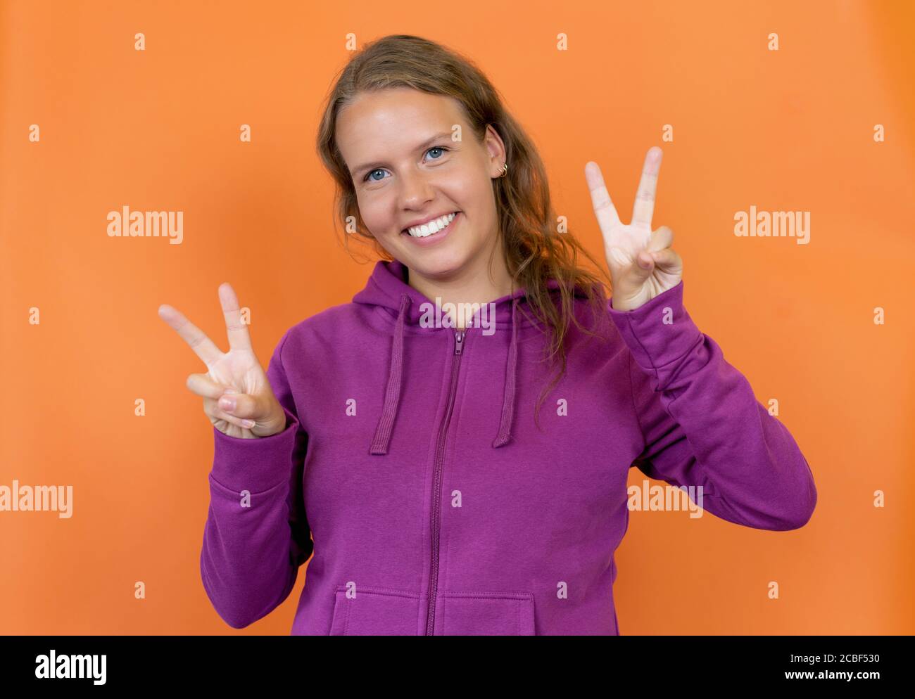 Blond german young adult woman with hoody showing victory gesture isolated on orange background Stock Photo