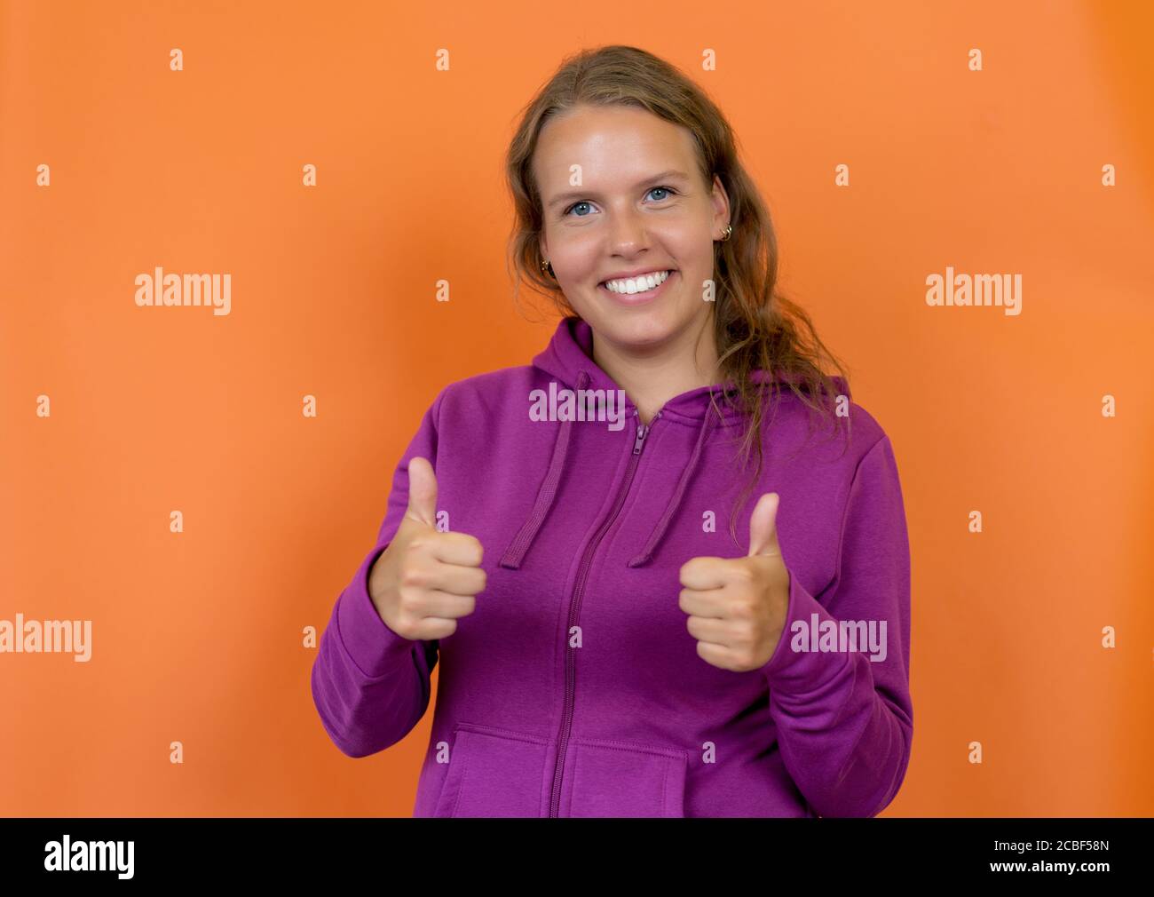 Funny blond german young adult woman with hoody showing thumbs up isolated on orange background Stock Photo