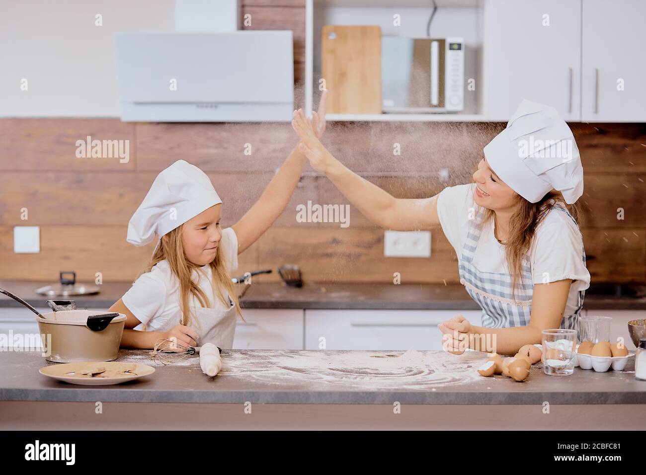 little daughter helping mother to bake, prepare dough. mom and kid dressed in aprons, clapping hands with each other Stock Photo