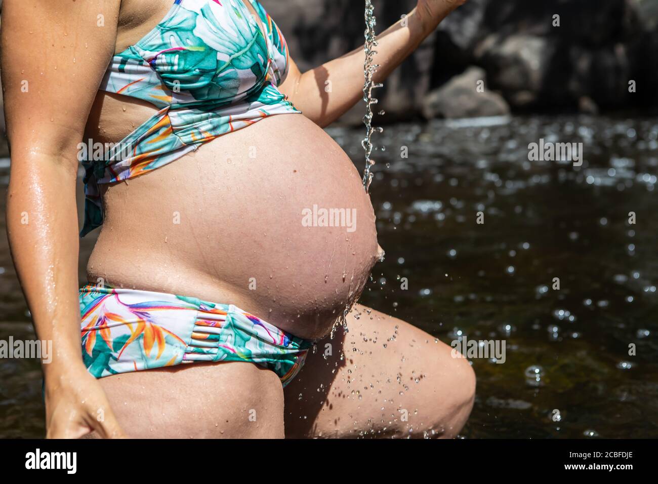 Young, beautiful pregnant woman in a bathing suit playing with water in a  mountain river and trickling some water from above on her big belly Stock  Photo - Alamy