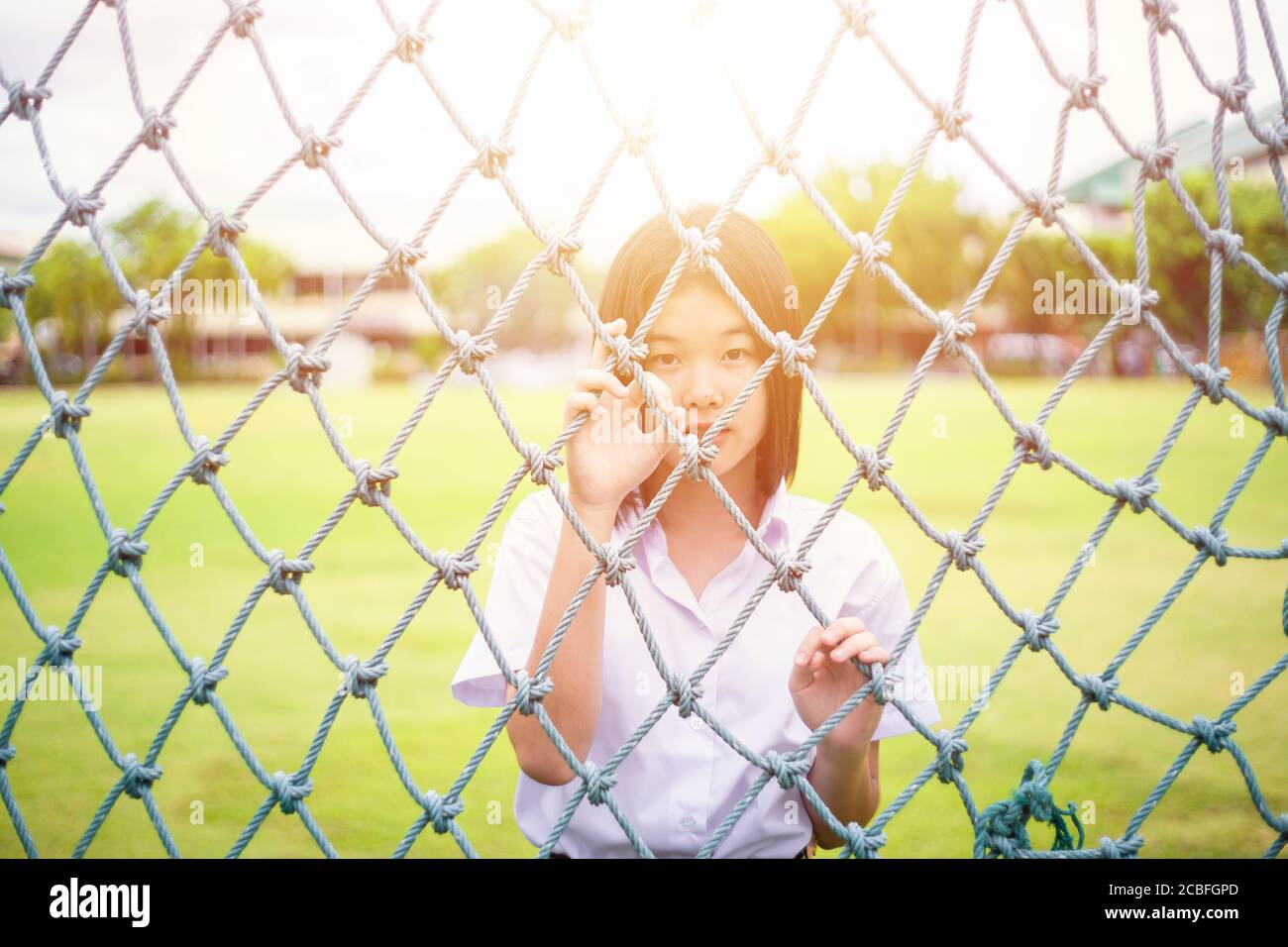 Cute portrait of innocent Asian girl teen student stand behind the rope net looking camera with sunshine Stock Photo