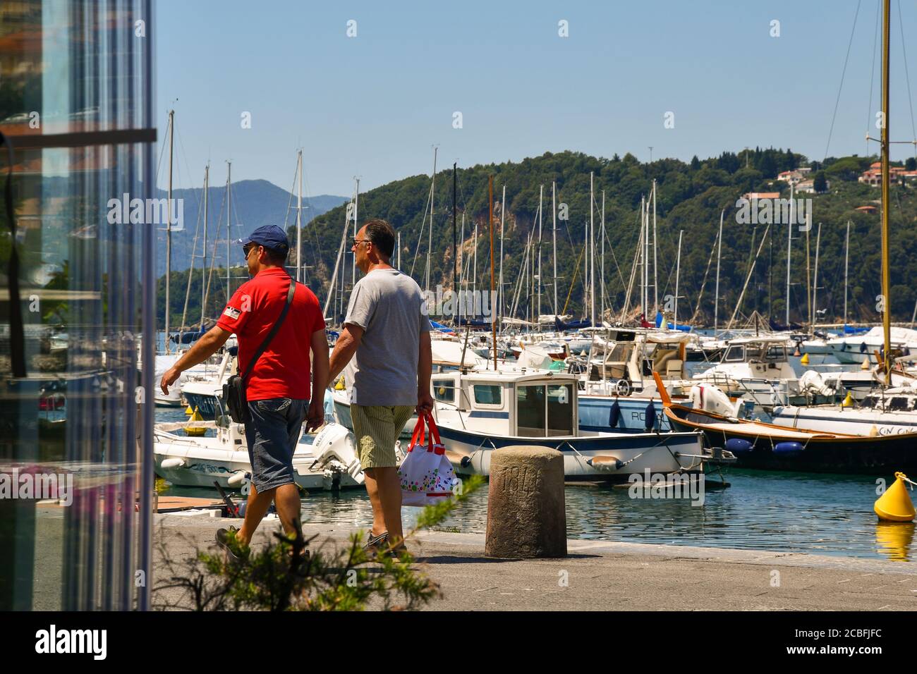 Two men walking on the quayside of the harbor with moored fishing and sail boats and the coast in the background in summer, Lerici, La Spezia, Italy Stock Photo