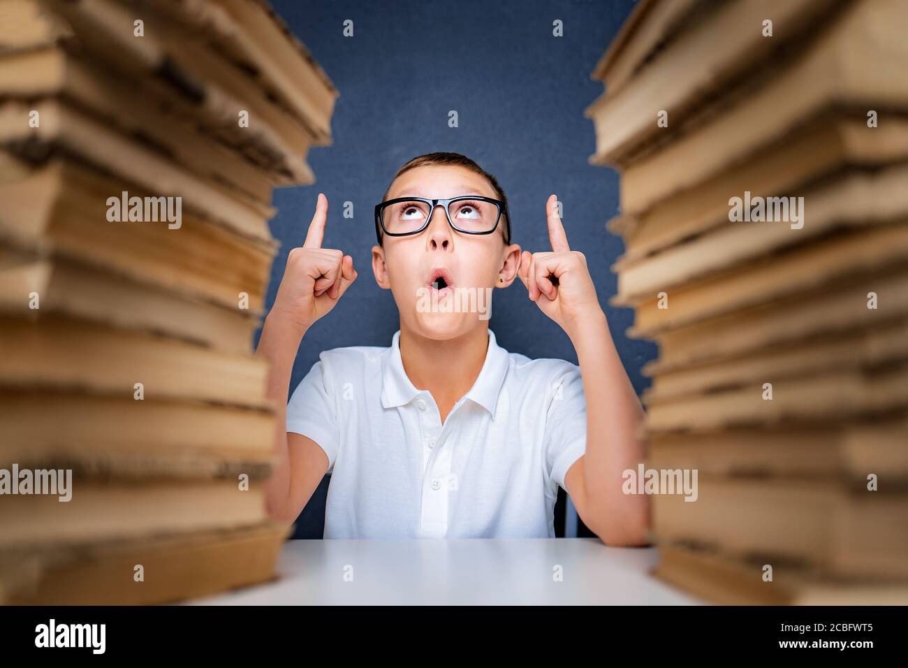 Smart boy in glasses sitting between two piles of books and look up