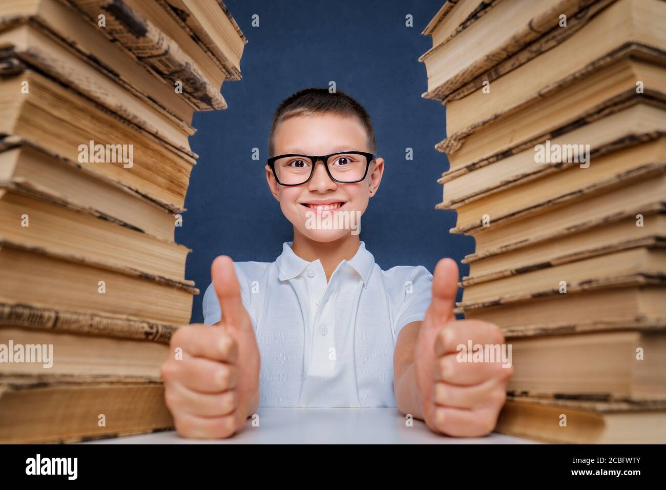 Happy smart boy in glasses sitting between two piles of books and look