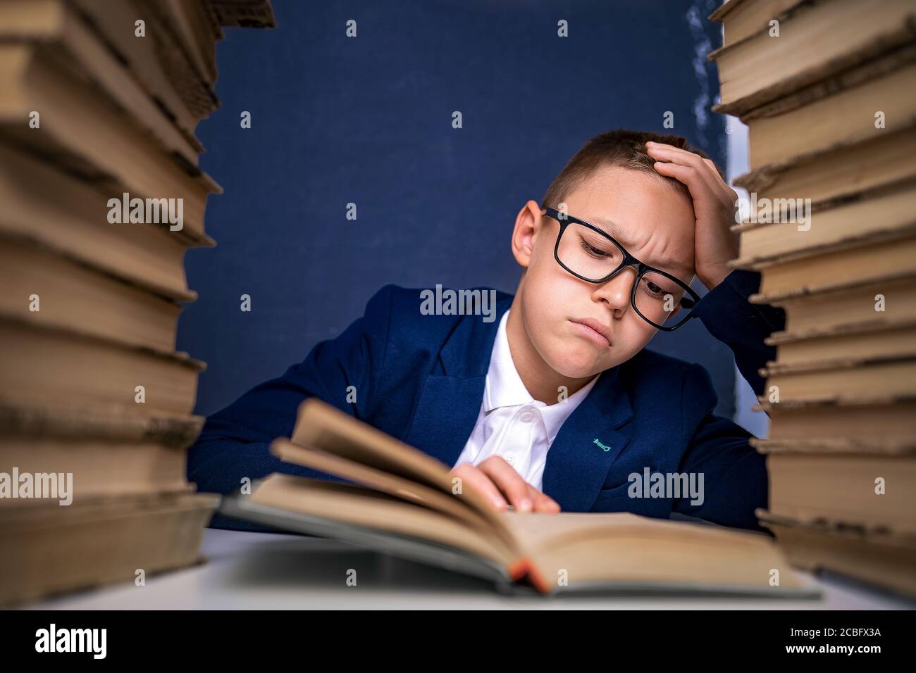 Smart boy in glasses sitting between two piles of books and read book thoughtfully. Stock Photo