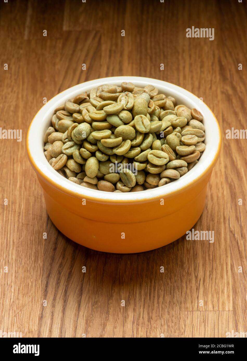 Green coffee beans in orange pot on wooden surface. Raw state before roasting. Stock Photo
