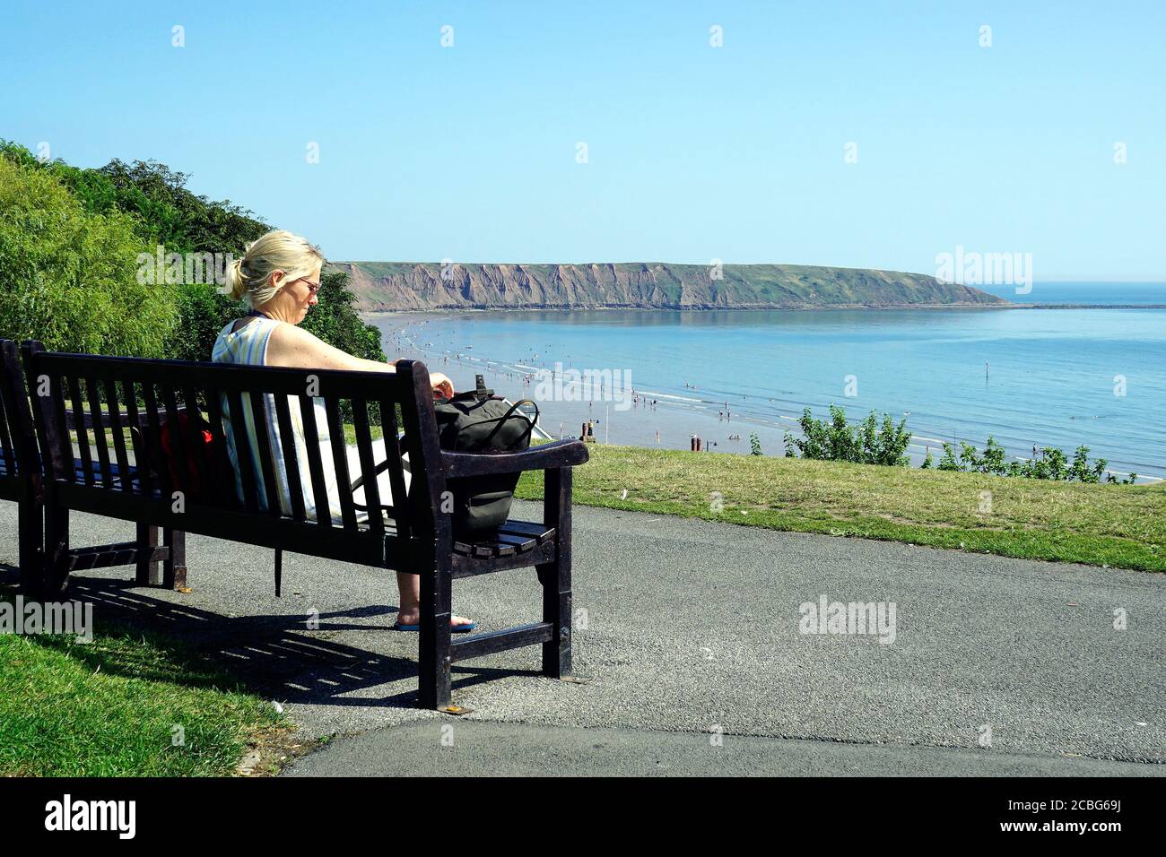 Filey, Yorkshire, UK.August 07, 2020.  Young lady relaxing on a seat at Crescent gardens overlooking the beach and bay at Filey in Yorkshire, UK. Stock Photo