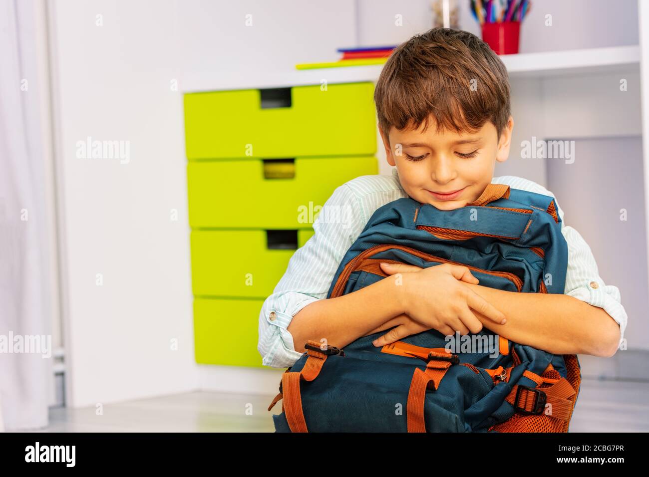 Close portrait of a smiling calm little boy sit in his room with positive expression holding school rucksack Stock Photo