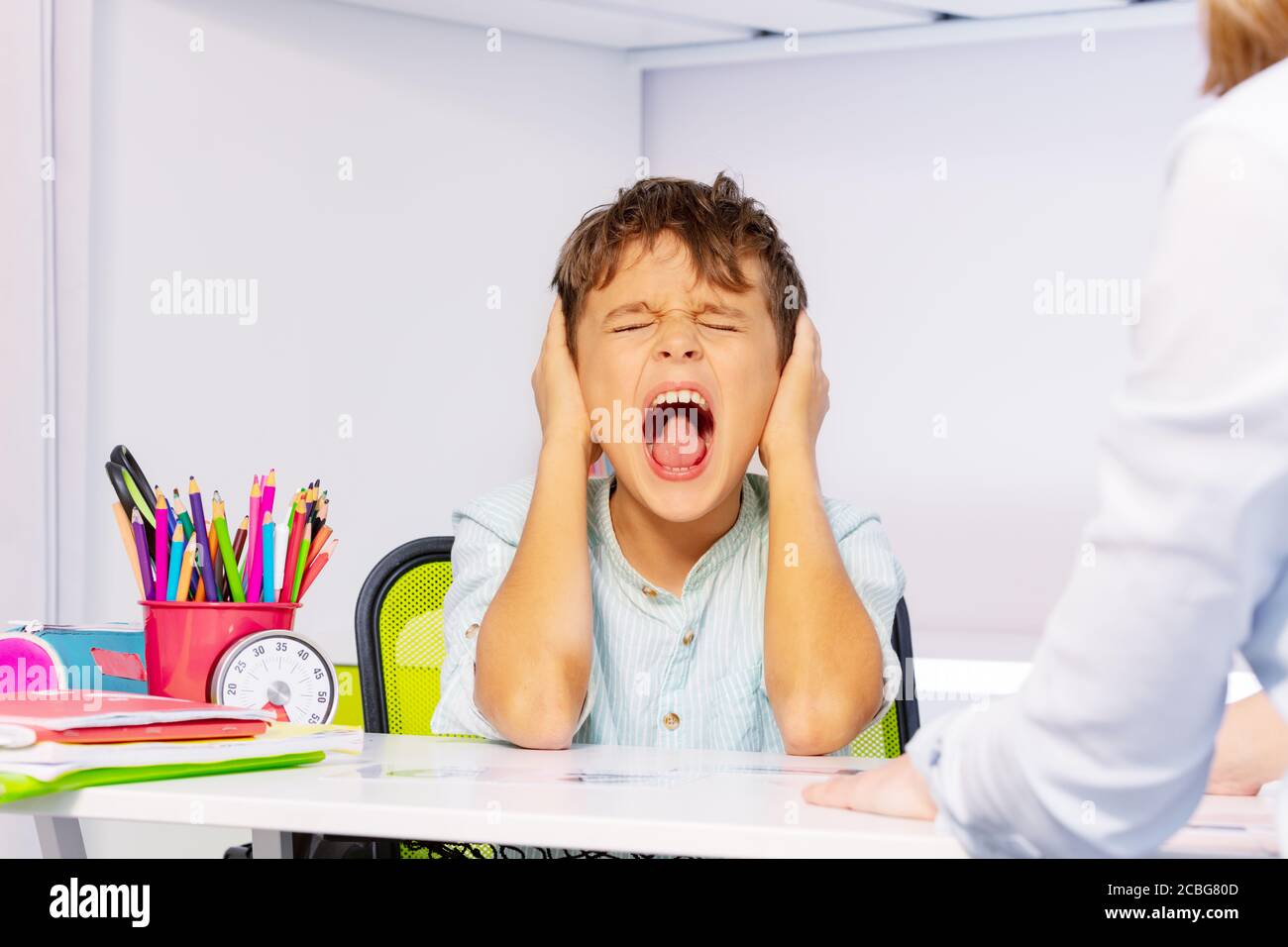 Boy with autism disorder scream and close ears in pain during ABA development therapy sitting by the table in class, sensory problem Stock Photo