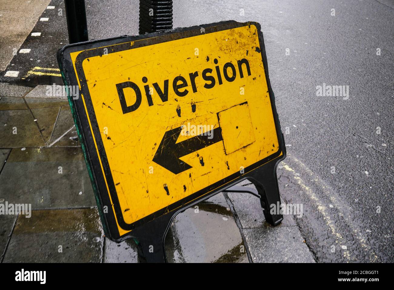 Yellow diversion sign with arrow pointing left on wet asphalt road. Stock Photo