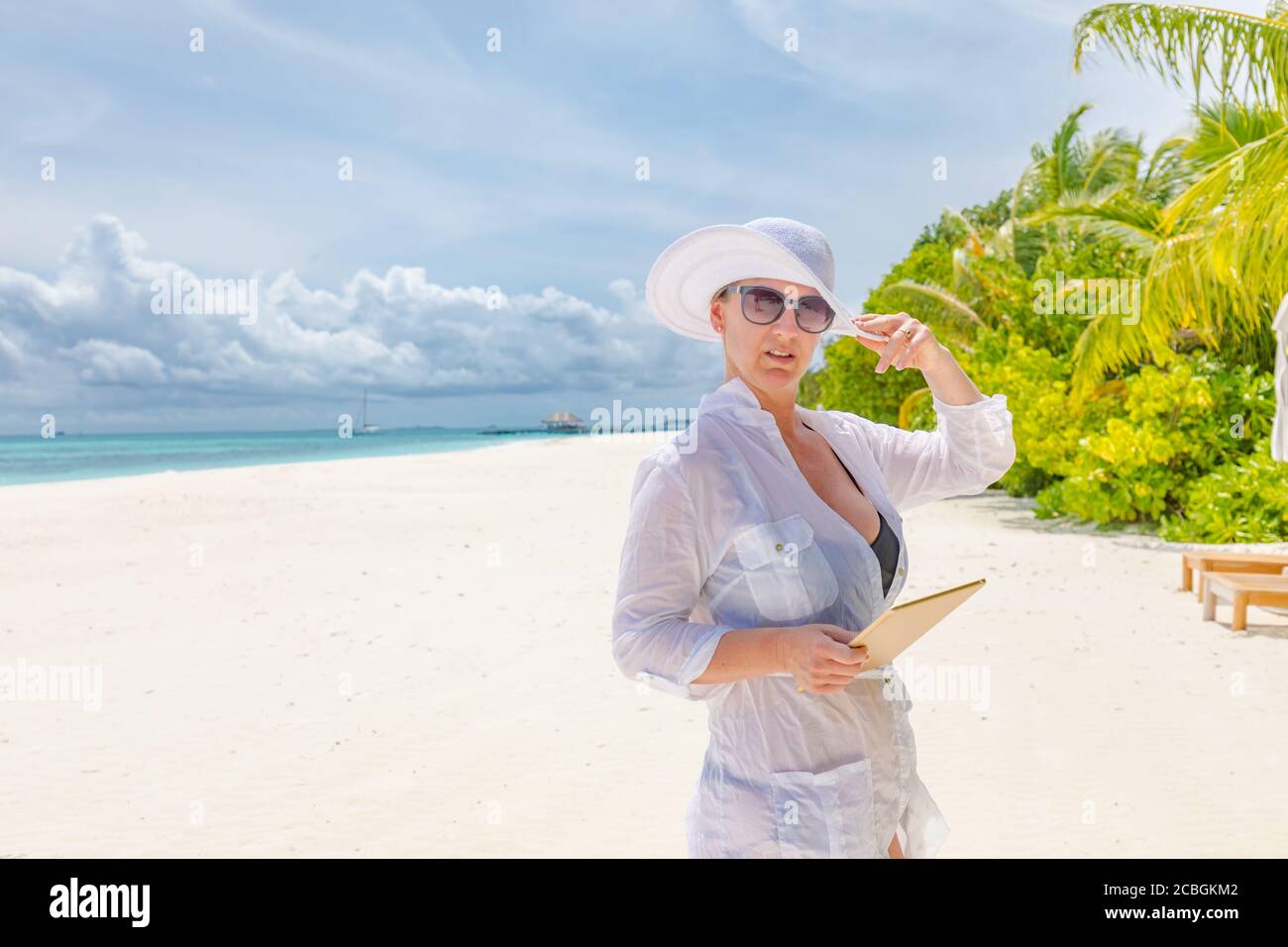 Woman using tablet computer on vacation in luxury resort, sunglasses and white dress with hat. Relaxation recreation and chaise lounge, leisure travel Stock Photo