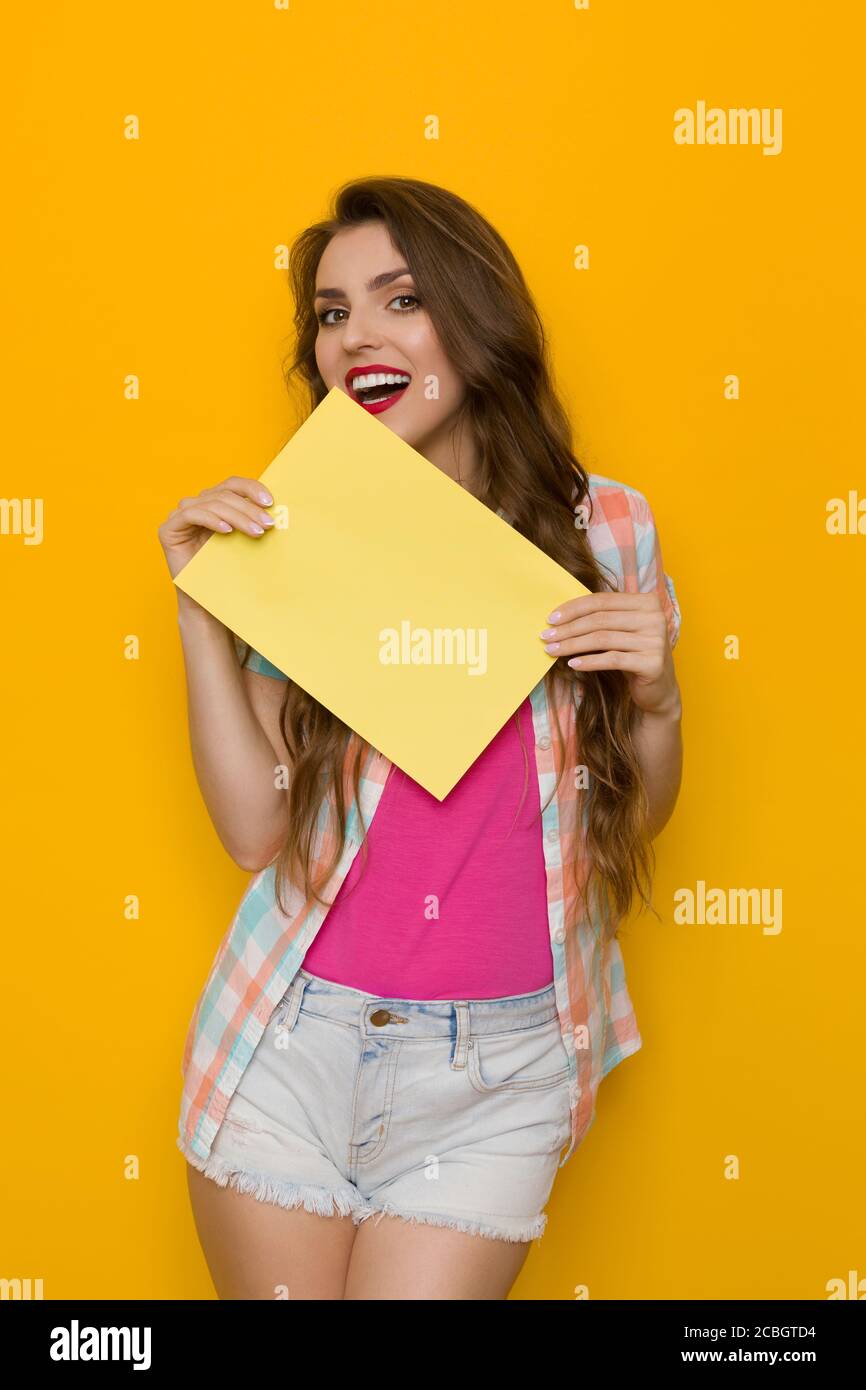 Happt beautiful young woman is holding yellow card and trying to bite it. Front view. Three quarter length studio shot on yellow background. Stock Photo