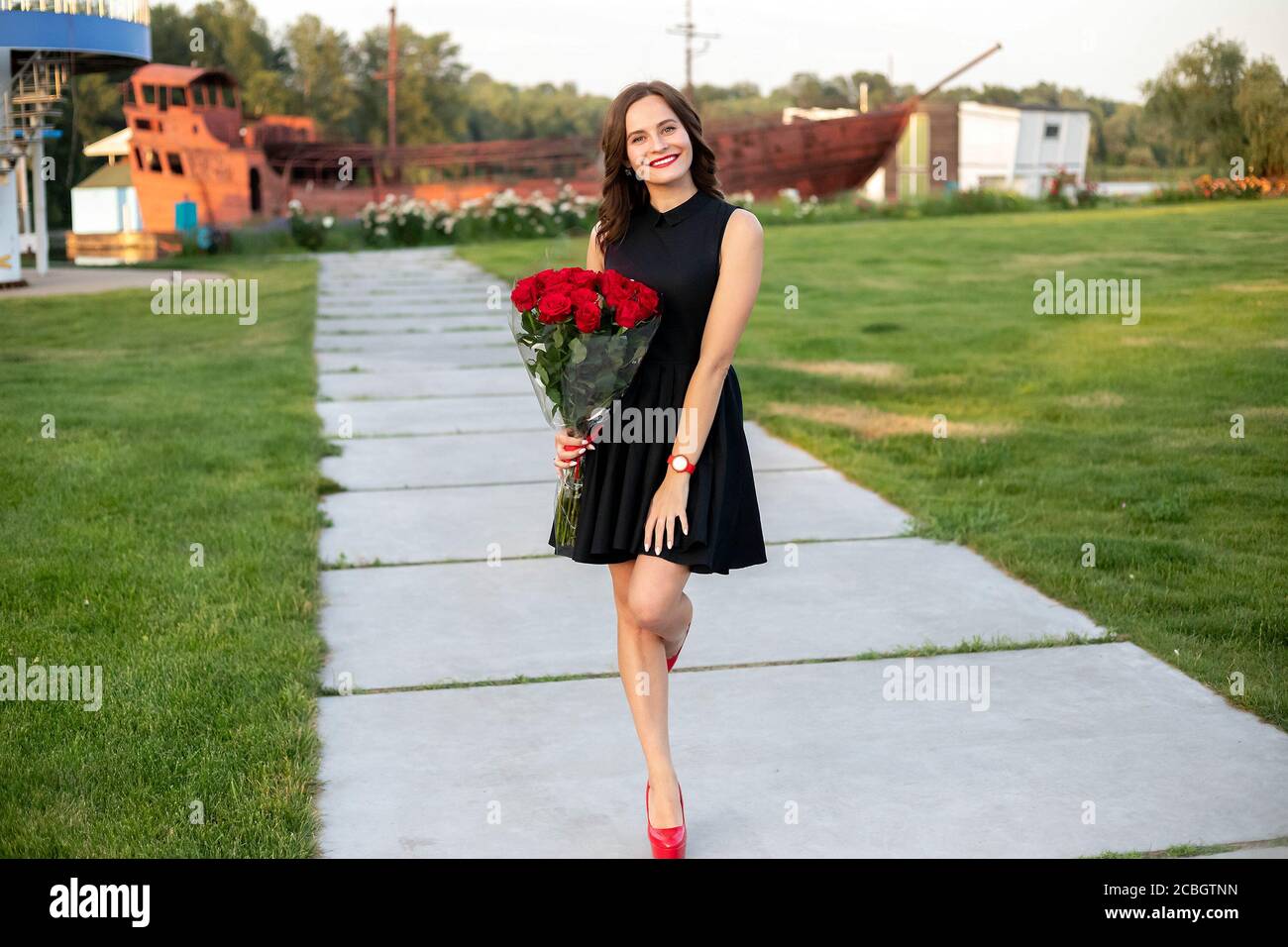 Attractive birthday girl with brown hair in little black dress smiling and holding a bouquet of red roses on background of wooden boat at the river co Stock Photo