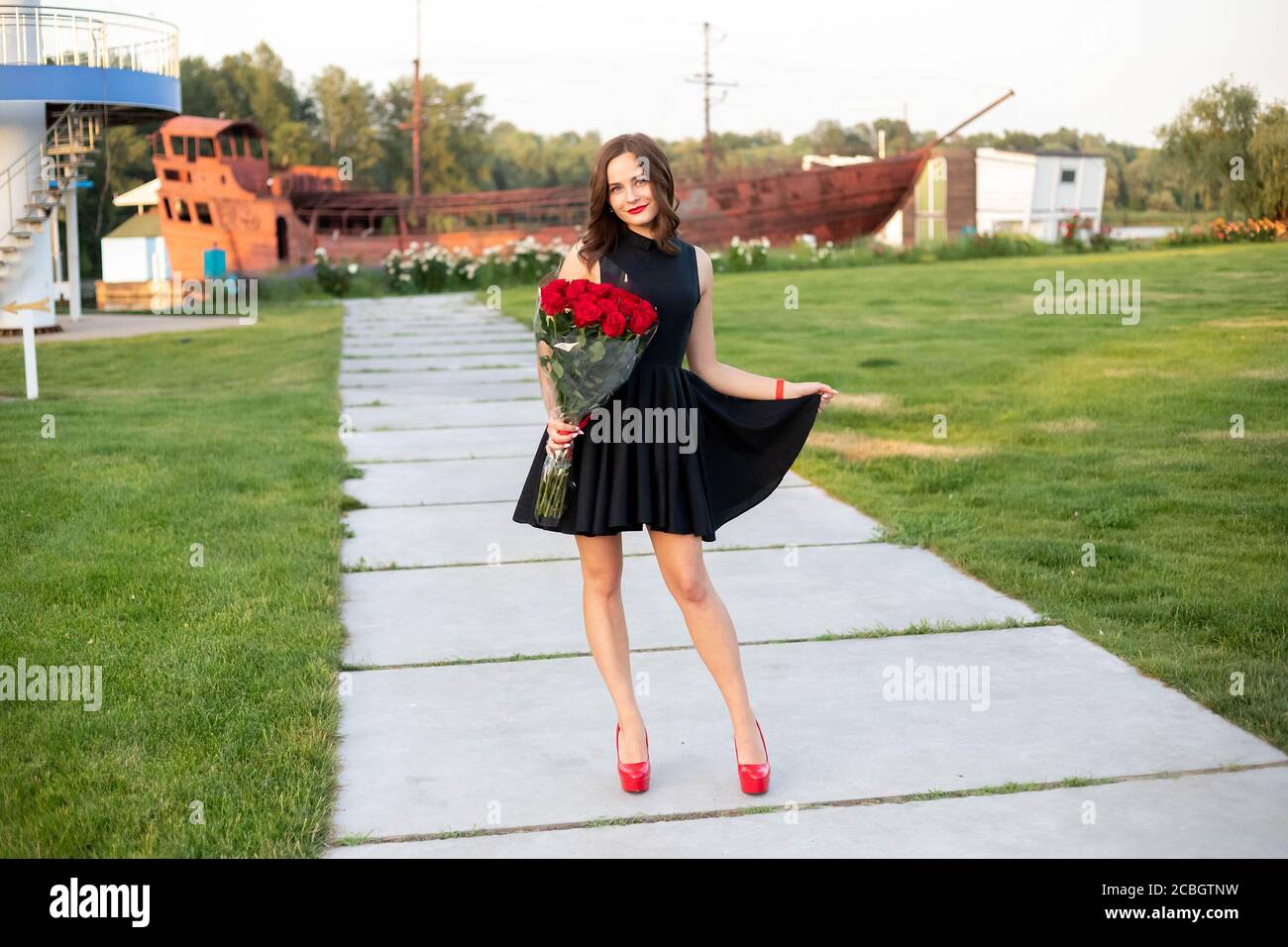 Attractive young woman with brown hair in little black dress posing, smiling and holding a bouquet of red roses on background of wooden boat at the ri Stock Photo
