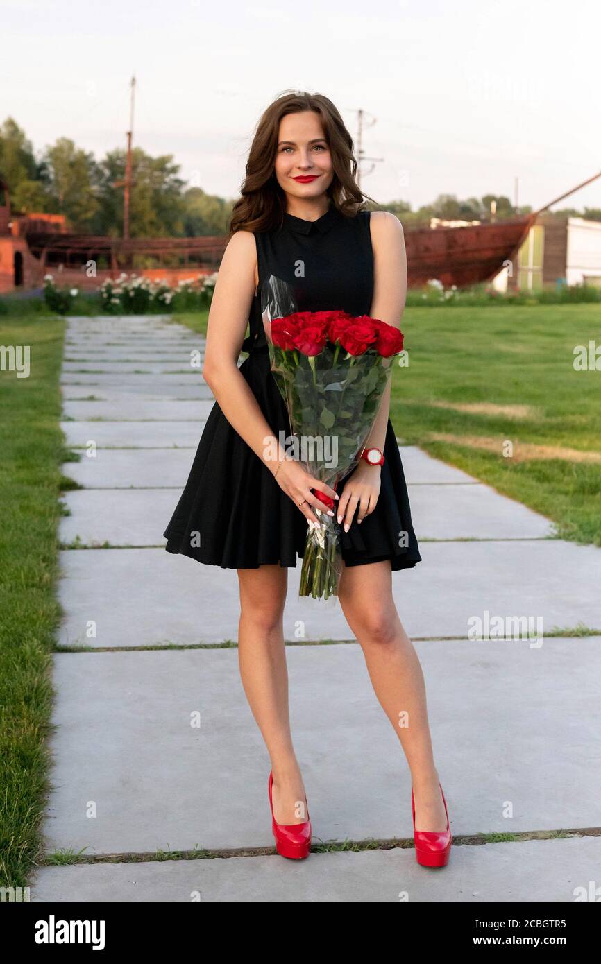 Pretty birthday girl with brown hair in little black dress posing, smiling and holding a bouquet of red roses on background of wooden boat at the rive Stock Photo