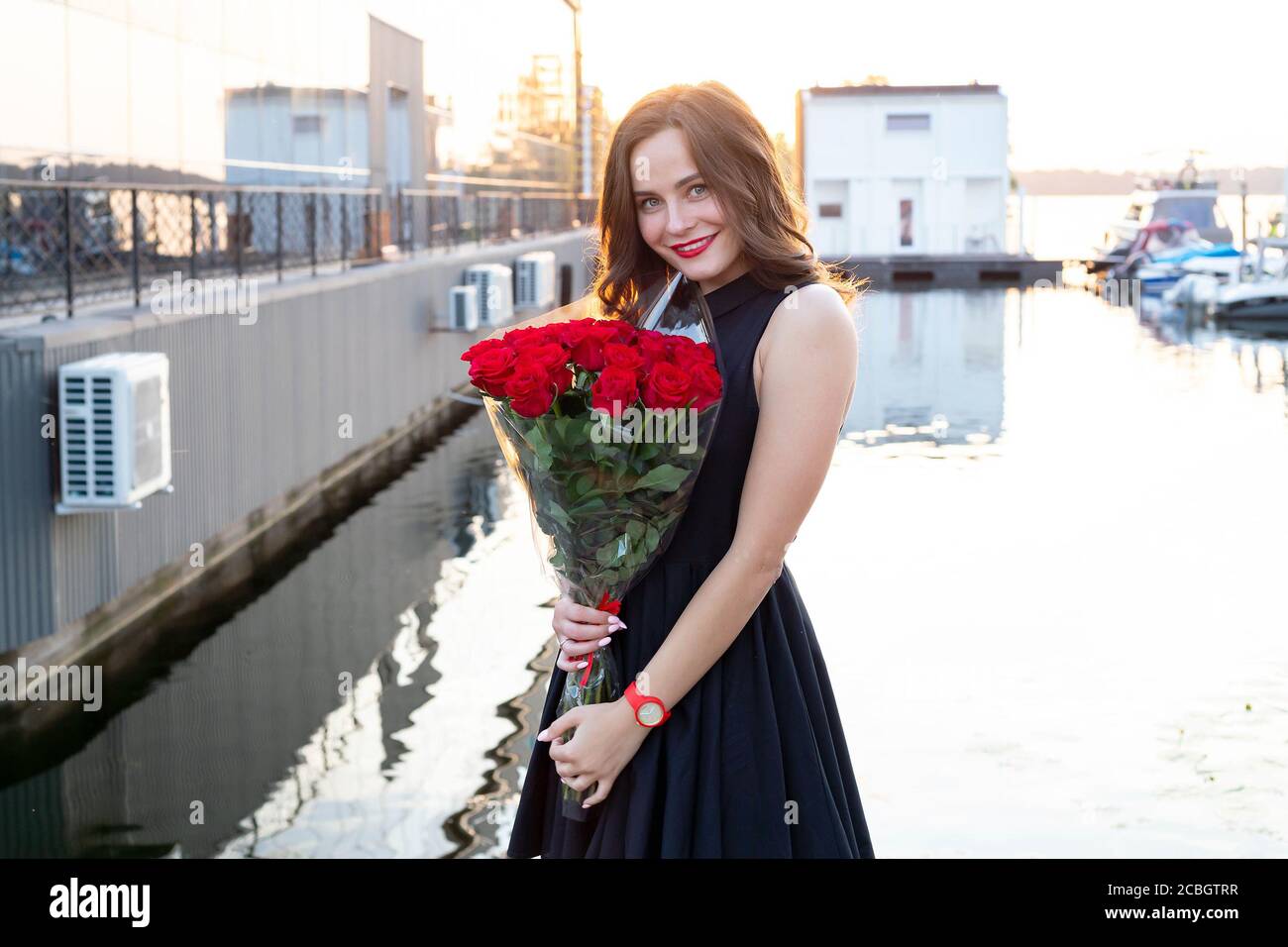 Portrait of attractive young woman with brown hair in little black dress smiling and holding a bouquet of red roses on background of the restaurant at Stock Photo