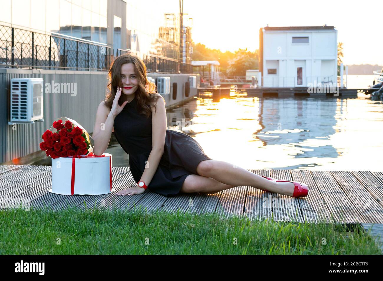 Attractive birthday girl with brown hair in little black dress sitting on the wooden footpath at sunset. Round box with birthday cake wrapped with red Stock Photo