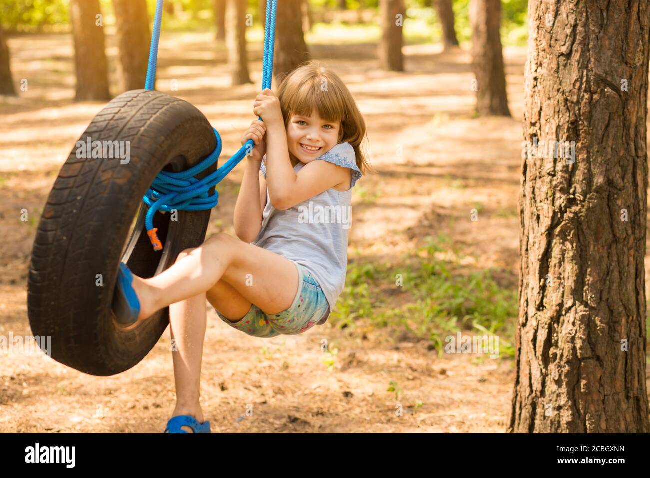 Happy active child girl playing on swing wheel in forest on sunny summer day. Summer outdoors activity for kids. Preschool child having fun and swingi Stock Photo