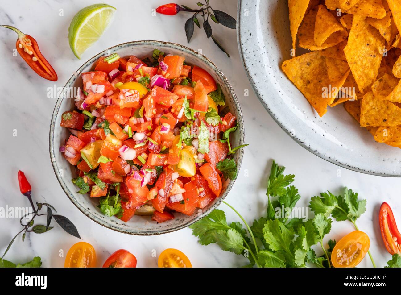 Tex Mex Pico de Gallo tomato salsa with nacho tortilla chips. The bowl is seen from above, flat lay, and there are ingredients scattered on the table: Stock Photo