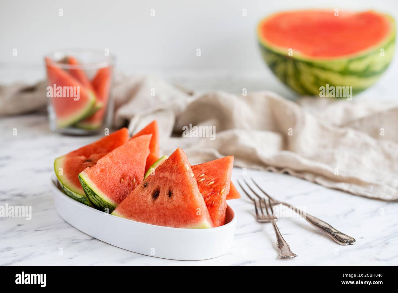 A bowl of watermelon slices with forks on the table. There is a halved watermelon in the background, a glass with some more pieces of melon and a line Stock Photo