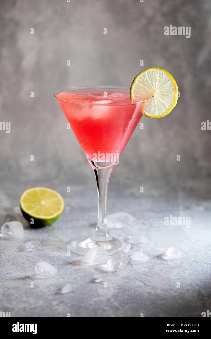 Cosmopolitan cocktail with lime. On a gray background with ice on the table, and there is a halved lime next to the glass. The fresh drink is garnised Stock Photo