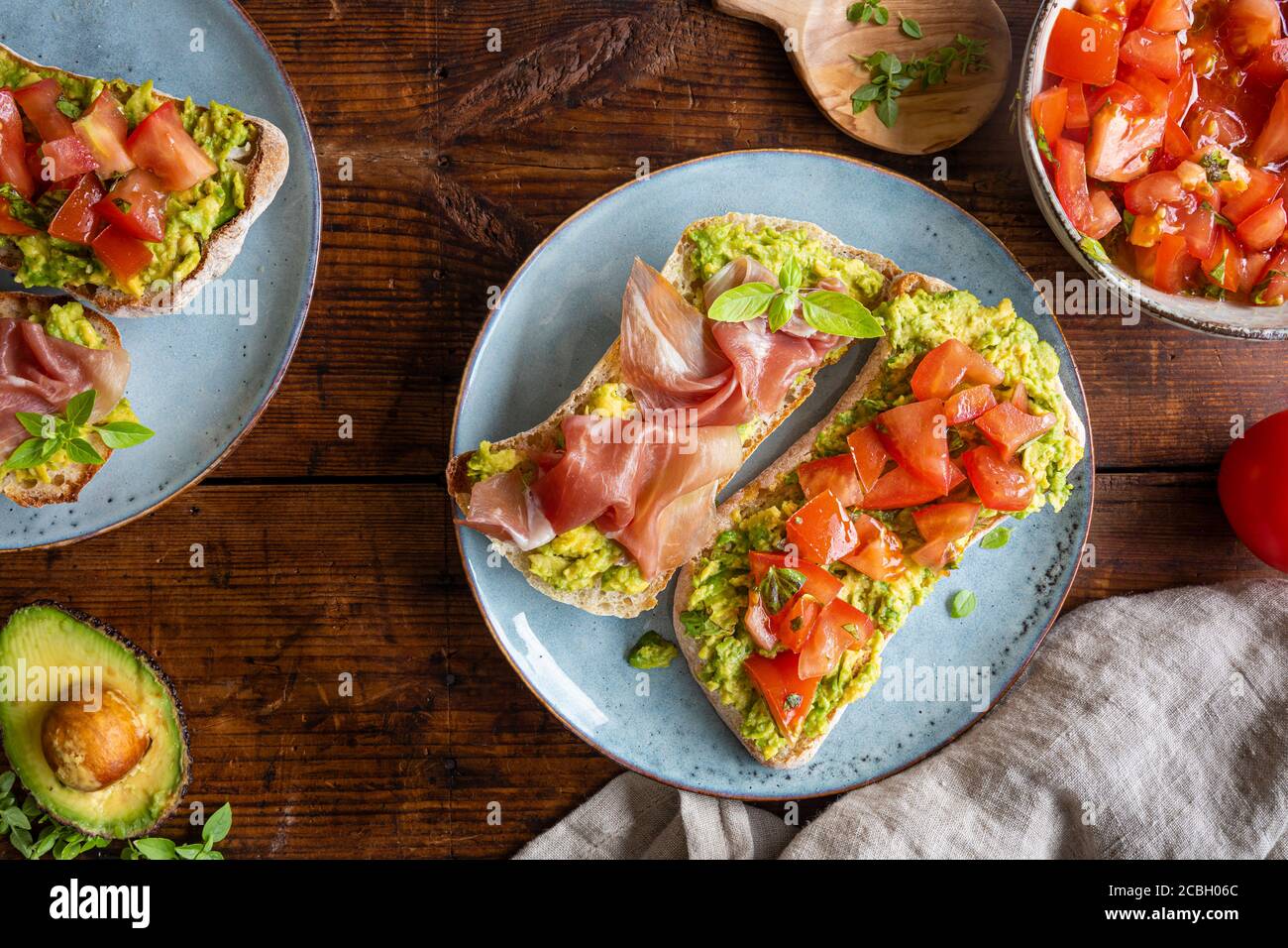 Avocado spread with chopped tomatoes and herbs. Parma prosciutto ham. Healthy breakfast seen from above. The food is on beautiful plates on a dark rus Stock Photo