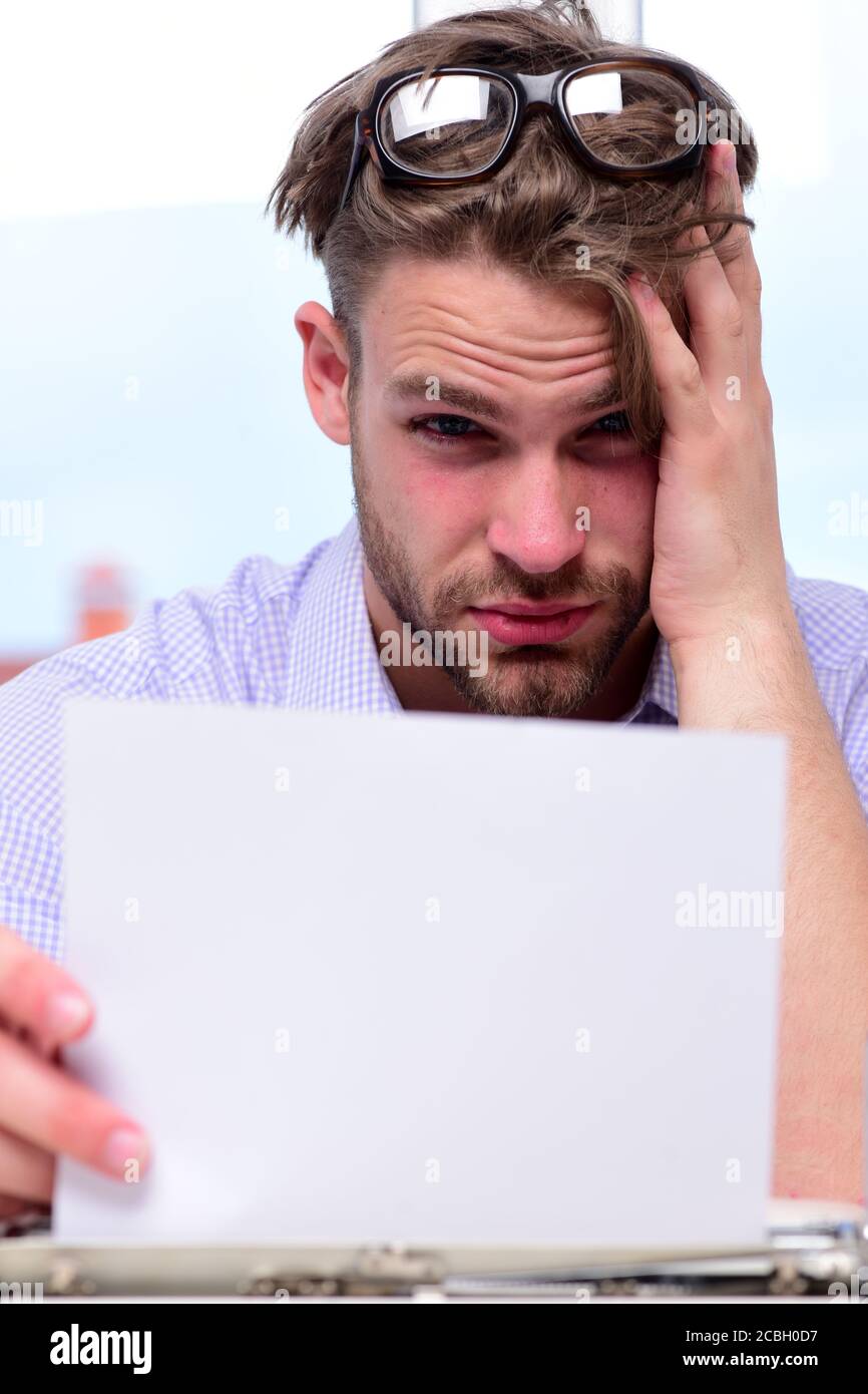 Man with thoughtful or troubled face at work. Writer holding white paper.  Handsome young author or editor with glasses on head looking for idea for  story on defocused background. Work and art