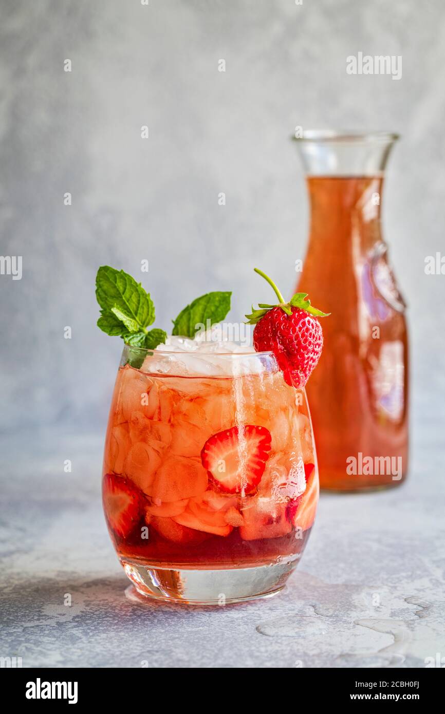 A red drink with ice and sliced strawberries, garnished with mint and a fresh strawberry., and a carafe of the same liquid in a the background.  The d Stock Photo